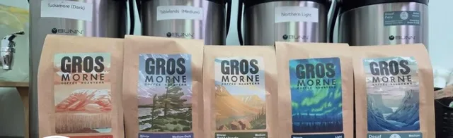 Five Gros Morne Coffee Roasters whole-bean bags in front of four labelled BUNN brewing urns at The Mill Coffee Company